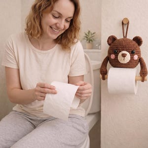 May include: A brown crocheted bear toilet paper holder featuring a round head with black eyes, a cream-colored muzzle, and pink cheeks. The bear has two arms extending downward to hold a wooden dowel that supports a roll of white toilet paper.