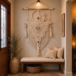 May include: A large off-white macrame wall hanging featuring a triangular design with intricate knot patterns, a central circular motif, and hanging tassels. The piece is suspended from a horizontal wooden rod with a cord and wooden beads.