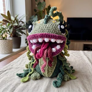 May include: A crocheted monster plush toy featuring a textured green body, a wide mouth with white teeth, and a dark pink interior. It has long, curly green tendrils, dark pink tongue-like appendages, and large eyes with long black eyelashes.