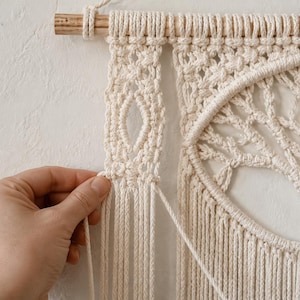 Pu&ograve; includere: Un arazzo da parete in macram&egrave; color crema con un design dell'albero della vita sospeso a un bastone di legno. L'intricato lavoro a corda annodata include un pannello verticale con un motivo a rombi aperto e una cornice circolare.