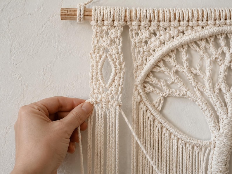 Pu&ograve; includere: Un arazzo da parete in macram&egrave; color crema con un design dell'albero della vita sospeso a un bastone di legno. L'intricato lavoro a corda annodata include un pannello verticale con un motivo a rombi aperto e una cornice circolare.