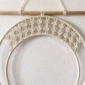 Pu&ograve; includere: Una decorazione da parete con un design circolare in macram&egrave; realizzato in corda di cotone color bianco sporco. L'oggetto &egrave; appeso a un bastone di legno orizzontale tramite un cappio, mostrando un motivo a nodi a griglia nel telaio superiore.