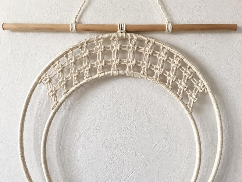 Pu&ograve; includere: Una decorazione da parete con un design circolare in macram&egrave; realizzato in corda di cotone color bianco sporco. L'oggetto &egrave; appeso a un bastone di legno orizzontale tramite un cappio, mostrando un motivo a nodi a griglia nel telaio superiore.