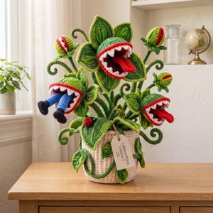 Könnte beinhalten: Eine gehäkelte fleischfressende Pflanzenskulptur in einem beigen Topf mit einem Etikett mit der Aufschrift CARNIVOROUS CROCHET CO. Die Pflanze hat grüne Blätter, rotlippige Münder mit weißen Zähnen, rote Zungen, einen Marienkäfer und blaue Beine.