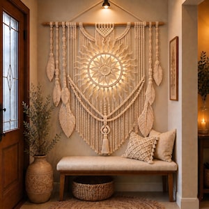Può includere: Un grande arazzo da parete in macramè color crema con un design centrale a raggiera solare e illuminazione calda integrata, sospeso a un bastone di legno. L'opera presenta complessi motivi a nodi, lunghe frange e dettagli decorativi a forma di piuma.