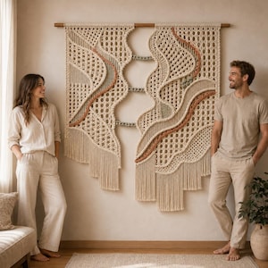 Puede incluir: Un tapiz de macramé grande e intrincado colgado de una varilla de madera. La pieza presenta cordón color crema tejido en complejos patrones geométricos, acentuado por líneas onduladas en terracota y verde salvia, con flecos largos abajo.