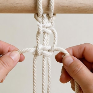 Pu&ograve; includere: Un progetto in corda macram&egrave; beige chiaro caratterizzato da una serie di nodi quadrati legati attorno a un bastoncino di legno liscio. La spessa corda di cotone ritorto &egrave; disposta in un motivo verticale con due estremit&agrave; libere tirate verso.