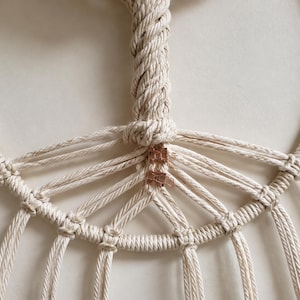 Pu&ograve; includere: Un arazzo in macram&egrave; realizzato con corda di cotone color bianco sporco annodata attorno a una cornice circolare. Il design presenta una sezione centrale verticale intrecciata che si dirama in diversi cordoni radianti, fissati con clip color rame.