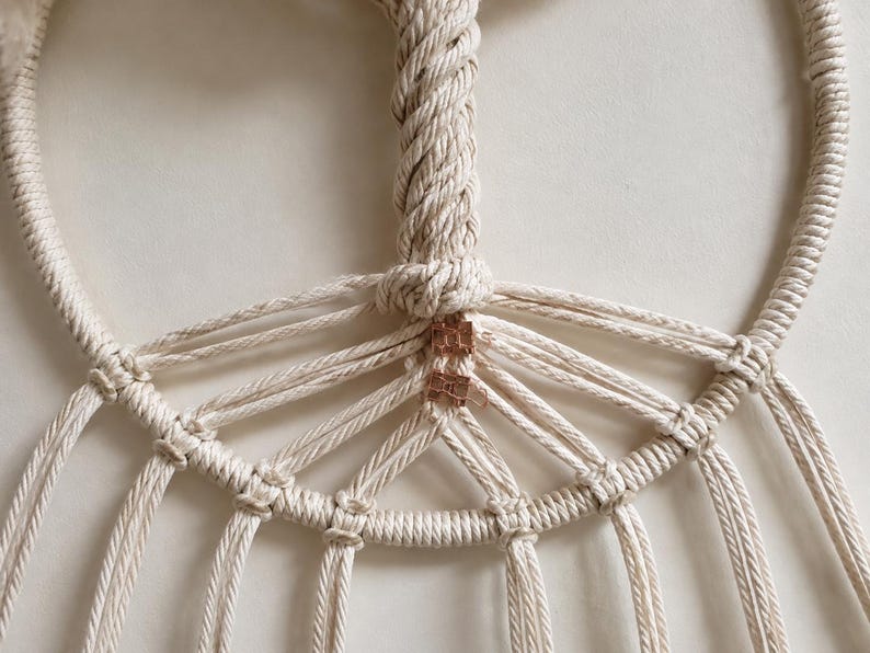 Pu&ograve; includere: Un arazzo in macram&egrave; realizzato con corda di cotone color bianco sporco annodata attorno a una cornice circolare. Il design presenta una sezione centrale verticale intrecciata che si dirama in diversi cordoni radianti, fissati con clip color rame.