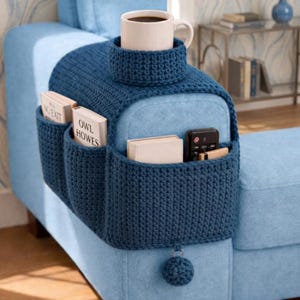 May include: A dark blue crocheted sofa armrest organizer featuring a built-in cup holder, multiple storage pockets containing books and a remote control, and a decorative crocheted pom-pom hanging from the bottom edge of the fabric.