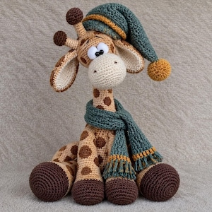 Könnte beinhalten: Ein gehäkeltes Giraffen-Stofftier mit beigem Körper und braunen Flecken, dunkelbraunen Hufen und cremefarbener Schnauze. Die Giraffe trägt einen türkisfarbenen Strickschal mit goldenen Fransen und eine passende Schlafmütze mit goldenem Bommel.