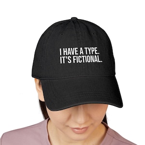 May include: A black baseball cap featuring the white embroidered text I HAVE A TYPE. IT'S FICTIONAL. The cap has a structured crown, a curved brim, and a button on top. The design is centered on the front panel of the hat in a clean, sans-serif font.