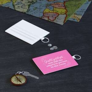 May include: A white card holder and a pink card holder, each with a silver metal keyring. The pink holder features the white text: Gaslit, gatekept, and still the smartest person in the room. Three small silver coins and a silver key are present.