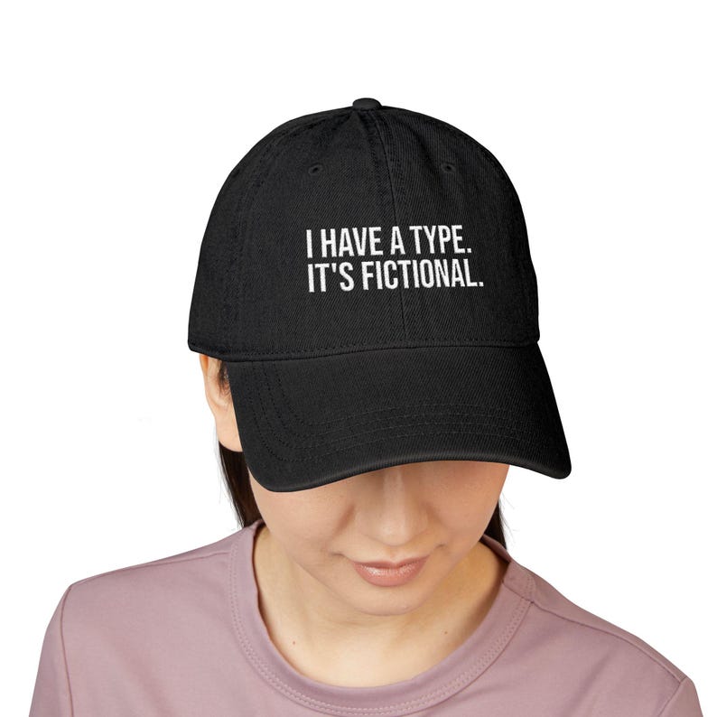May include: A black baseball cap featuring the white embroidered text I HAVE A TYPE. IT'S FICTIONAL. The cap has a structured crown, a curved brim, and a button on top. The design is centered on the front panel of the hat in a clean, sans-serif font.