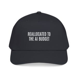 May include: A dark charcoal baseball cap featuring the white embroidered text REALLOCATED TO THE AI BUDGET centered on the front panel. The cap has a structured crown, a curved brim, and a button at the top center of the dome.