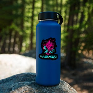 May include: A matte blue water bottle with a black screw-top lid. A sticker on the front features a stylized pink and cyan graphic of a samurai mask with horizontal glitch lines and the text SAMURAI printed in a bold, blocky, digital font.