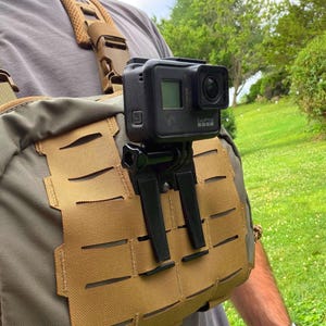May include: A black action camera mounted on a tan tactical chest rig. The camera is secured with a black plastic mount attached to the MOLLE webbing of the chest pack. The rig features adjustable brown shoulder straps with plastic buckles.