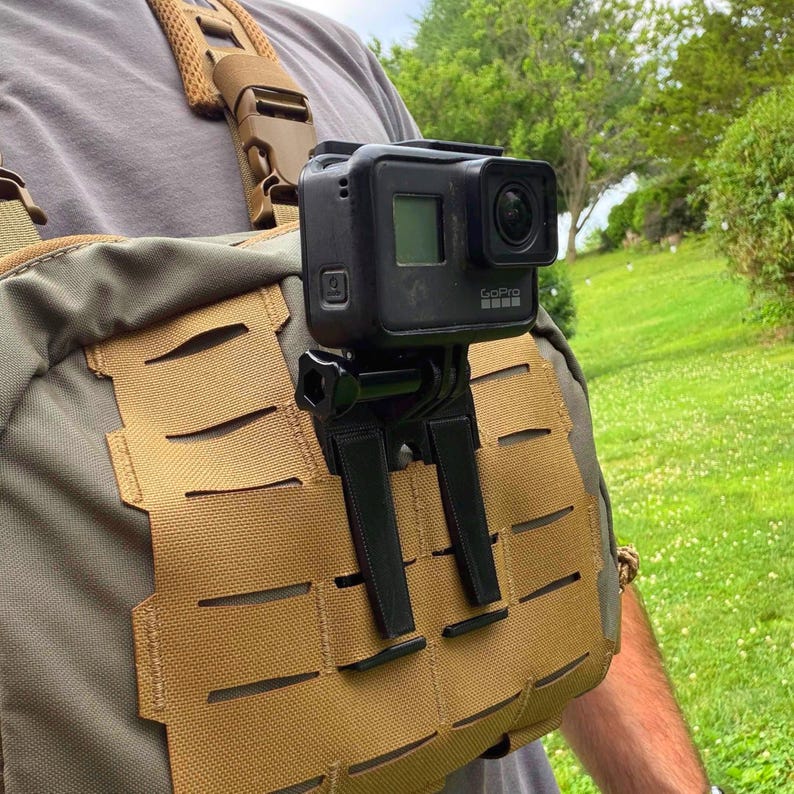 May include: A black action camera mounted on a tan tactical chest rig. The camera is secured with a black plastic mount attached to the MOLLE webbing of the chest pack. The rig features adjustable brown shoulder straps with plastic buckles.