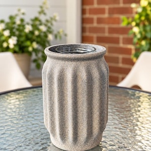 May include: A cylindrical vase with a textured, speckled gray stone-like finish and vertical fluted grooves. The vase features a wide, open top with a smooth, clear glass insert visible inside the center, creating a contrast in material and texture.