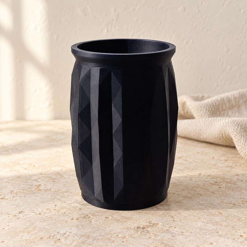 May include: A matte black cylindrical container featuring a geometric faceted exterior pattern. The vessel has a smooth, circular rim and a solid base, with vertical columns of triangular indentations creating a textured surface around the body.