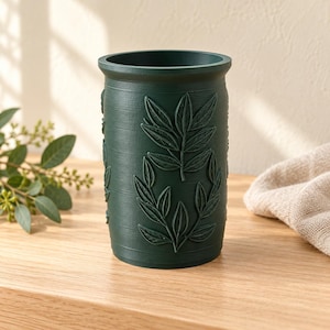 May include: A dark green cylindrical container featuring a textured surface with embossed leaf patterns. The vessel has a wide open top and a smooth rim, displaying a matte finish with vertical striations across the exterior body.