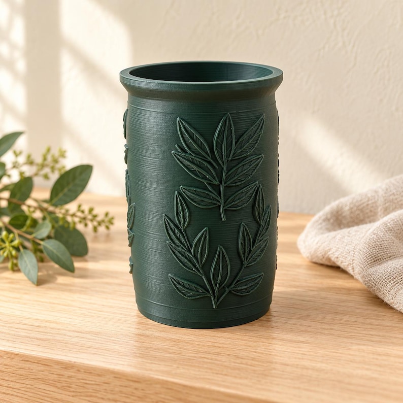 May include: A dark green cylindrical container featuring a textured surface with embossed leaf patterns. The vessel has a wide open top and a smooth rim, displaying a matte finish with vertical striations across the exterior body.