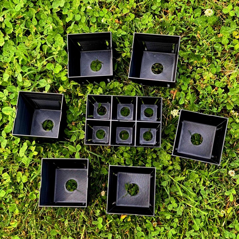 May include: A collection of black plastic square plant pots, including four individual larger pots and one connected tray containing six smaller square compartments, each featuring a circular drainage hole at the base of the container.