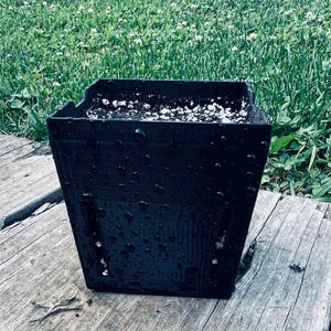 May include: A square black plastic plant pot with vertical drainage slots on the sides. The exterior surface is covered in water droplets, and the interior is filled with dark potting soil mixed with small white perlite particles.