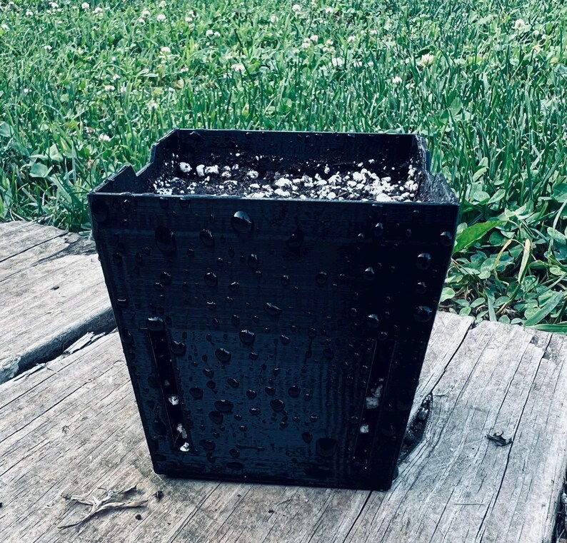 May include: A square black plastic plant pot with vertical drainage slots on the sides. The exterior surface is covered in water droplets, and the interior is filled with dark potting soil mixed with small white perlite particles.