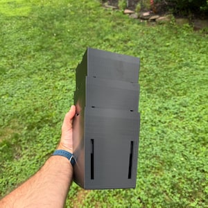 May include: A matte black, three-tiered, rectangular storage container with vertical slots on the sides. The object features a stepped, modular design with visible horizontal lines from the 3D printing process, constructed from a solid plastic material.