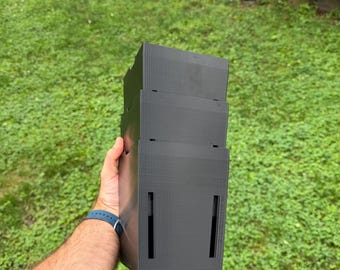 May include: A matte black, three-tiered, rectangular storage container with vertical slots on the sides. The object features a stepped, modular design with visible horizontal lines from the 3D printing process, constructed from a solid plastic material.
