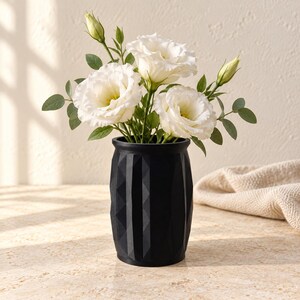 May include: A matte black vase with a geometric, faceted exterior surface. The vase contains a bouquet of white eustoma flowers with green leaves and several unopened flower buds, creating a contrast between the dark vessel and the light blooms.