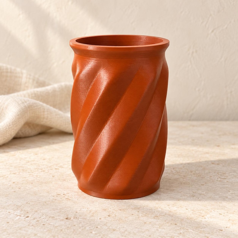 May include: A cylindrical terracotta-colored vessel featuring a distinct spiral or twisted pattern along its exterior surface. The object has a smooth, uniform finish with a wide, open top and a flat base, maintaining a consistent color throughout.