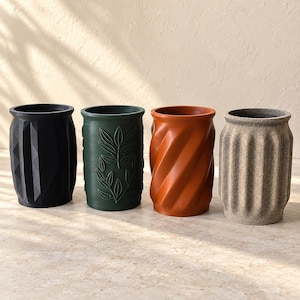 May include: Four cylindrical containers arranged in a row. The first is matte black with a geometric faceted surface. The second is dark green with embossed leaf patterns. The third is terracotta orange with a spiral twist. The fourth is beige with vertical fluted ridges.