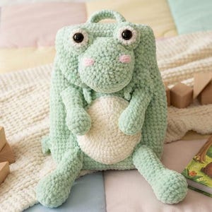 Puede incluir: Un juguete de peluche tejido a ganchillo de color verde claro con forma de dinosaurio, con una barriga redonda de color crema, ojos grandes y oscuros y mejillas rosadas. El juguete tiene un asa pequeña en la parte superior, brazos y piernas cortos.