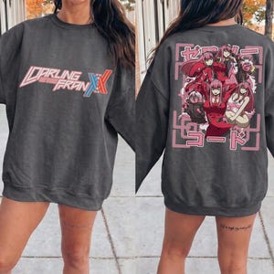 May include: A charcoal gray crewneck sweatshirt featuring the Darling in the Franxx logo in white and red on the front. The back displays a graphic print of the character Zero Two in various poses, surrounded by pink Japanese text and a square frame.