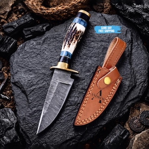 May include: A fixed-blade knife with a patterned Damascus steel blade, a brass guard, and a handle crafted from textured antler and blue resin. Beside it lies a tan leather sheath featuring a snap closure and the engraved name ANDREW.