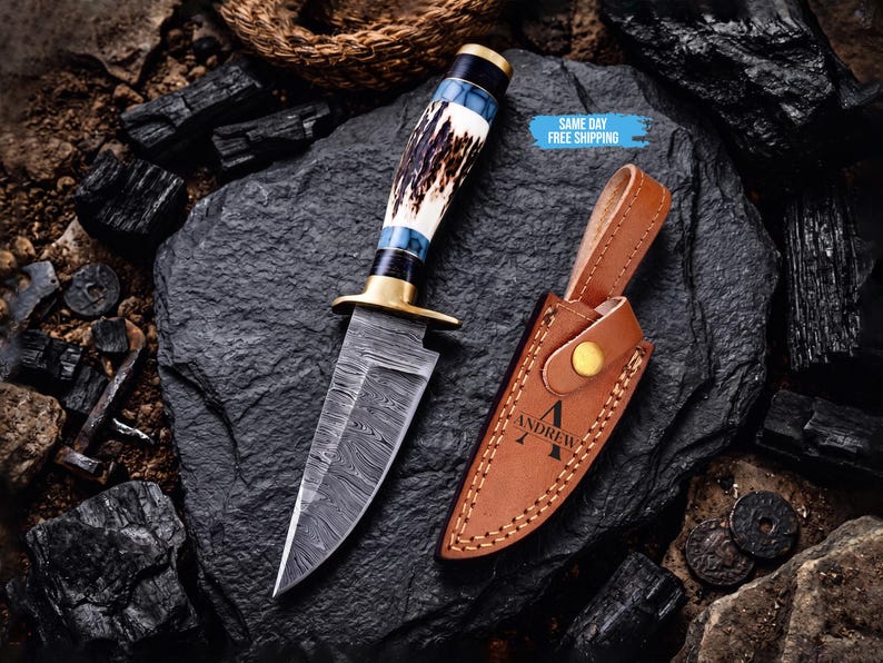 May include: A fixed-blade knife with a patterned Damascus steel blade, a brass guard, and a handle crafted from textured antler and blue resin. Beside it lies a tan leather sheath featuring a snap closure and the engraved name ANDREW.
