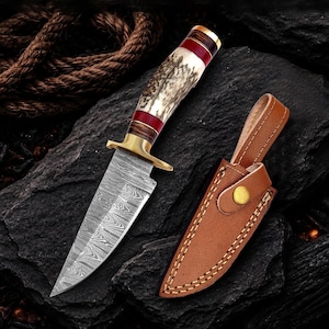 May include: A fixed-blade knife with a Damascus steel blade, a brass guard, and a handle crafted from textured antler with red and dark wood accents. The knife is accompanied by a tan leather sheath featuring visible stitching and a brass snap button.