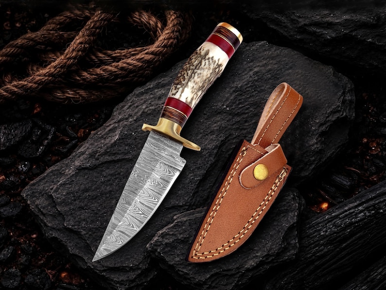 May include: A fixed-blade knife with a Damascus steel blade, a brass guard, and a handle crafted from textured antler with red and dark wood accents. The knife is accompanied by a tan leather sheath featuring visible stitching and a brass snap button.