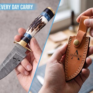 May include: A fixed-blade knife with a Damascus steel blade featuring the name ANDREW, a brass guard, and a handle made of antler and blue resin. Beside it, a tan leather sheath with a brass snap closure and the name ANDREW embossed on it.