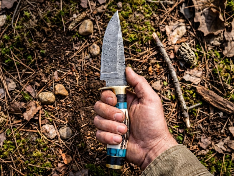 May include: A fixed-blade knife featuring a patterned Damascus steel blade, a brass guard, and a handle constructed from segments of dark wood, turquoise-colored stone, and textured antler, finished with a brass pommel at the base.