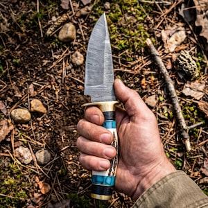 May include: A fixed-blade knife featuring a patterned Damascus steel blade, a brass guard, and a handle constructed from segments of dark wood, turquoise-coloured stone, and textured antler, finished with a brass pommel cap at the base.