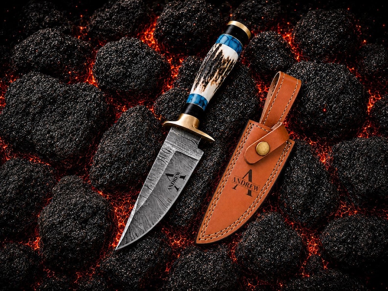 May include: A fixed-blade knife with a Damascus steel blade featuring the name Andrew, a brass guard, and a handle made of antler and blue resin. Beside it lies a tan leather sheath with a brass snap closure and the name Andrew embossed on it.