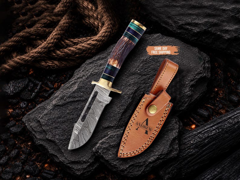 May include: A fixed-blade knife with a Damascus steel blade featuring a cutout, a brass guard, and a handle made of textured brown antler with teal and black spacers. Beside it lies a tan leather sheath embossed with the name ANDREW.