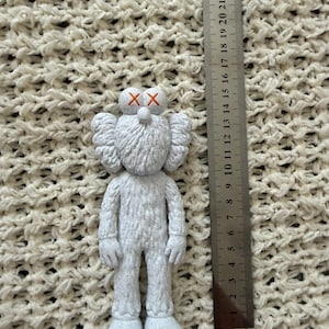 Puede incluir: Una figura KAWS blanca con superficie texturizada y s&iacute;mbolos X naranjas en los ojos, colocada junto a una regla met&aacute;lica que muestra que la figura mide aproximadamente 15 cm de altura. La figura tiene orejas grandes, cabeza redondeada y textura.
