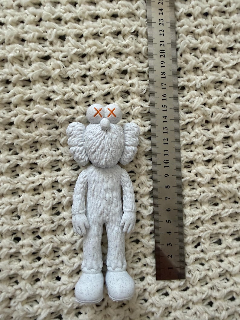Puede incluir: Una figura KAWS blanca con superficie texturizada y s&iacute;mbolos X naranjas en los ojos, colocada junto a una regla met&aacute;lica que muestra que la figura mide aproximadamente 15 cm de altura. La figura tiene orejas grandes, cabeza redondeada y textura.