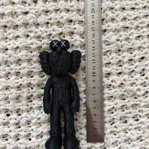 Puede incluir: Una figura KAWS negra con ojos en forma de X, colocada junto a una regla de metal que muestra que la figura mide aproximadamente 14,5 cent&iacute;metros de altura. La figura tiene una apariencia texturizada similar al pelaje, orejas redondeadas y pies grandes.