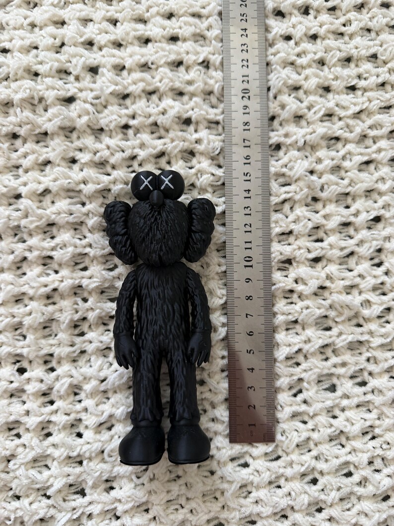 Puede incluir: Una figura KAWS negra con ojos en forma de X, colocada junto a una regla de metal que muestra que la figura mide aproximadamente 14,5 cent&iacute;metros de altura. La figura tiene una apariencia texturizada similar al pelaje, orejas redondeadas y pies grandes.