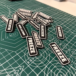 May include: A collection of rectangular black tokens with white borders and white text. The tokens feature the words FREEZE, BLOCKER, or ABILITY USED in a bold, sans-serif font. These small, flat game markers are scattered across a green surface.